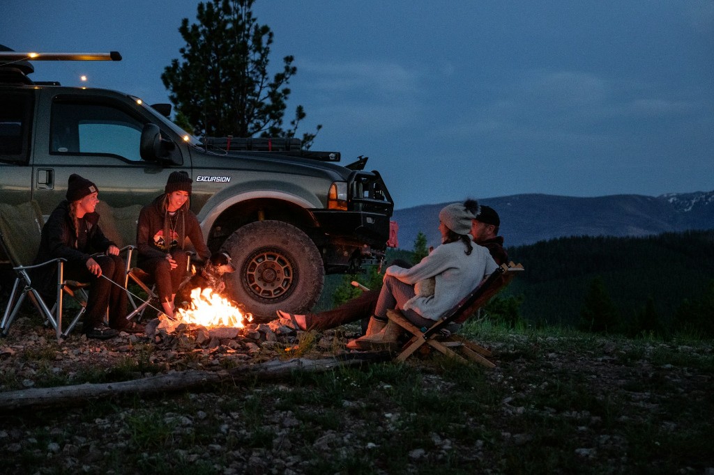 Group around a campfire beside a modified Ford Excursion at dusk in the mountains